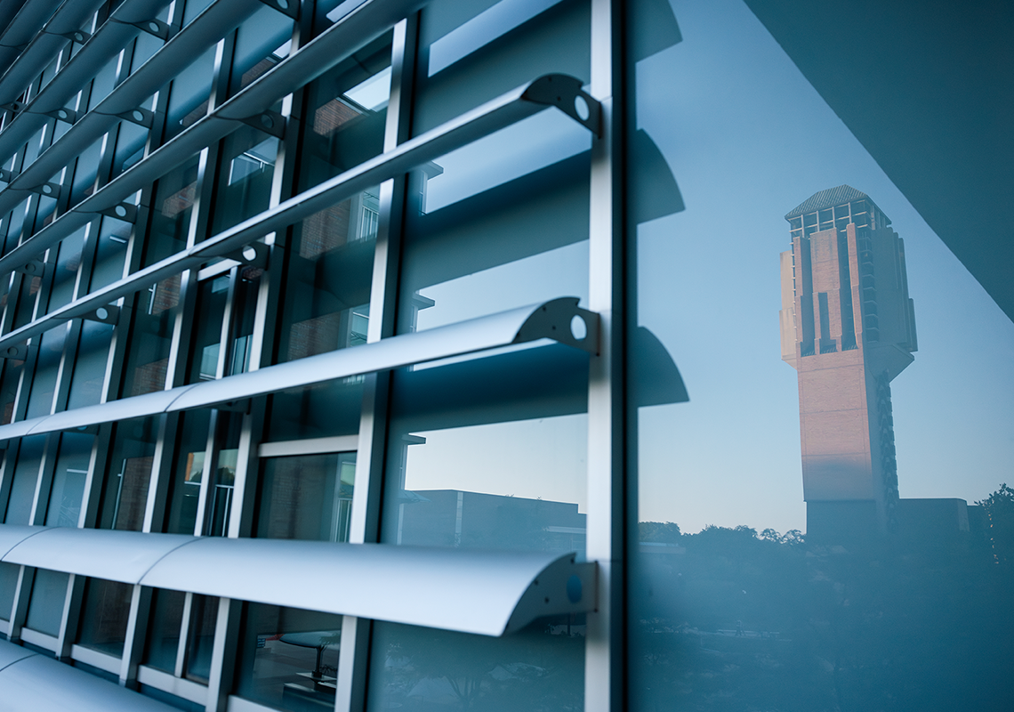 View of Lurie Bell Tower reflected in the glass facade of a modern campus building, framed by horizontal metal sunshades. The blue-tinted windows create a geometric pattern, contrasting with the warm brick tones of the tower in the reflection.
