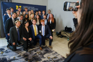 A group of professionally dressed individuals poses for a photo in front of a banner for the Tauber Institute for Global Operations at the University of Michigan, while someone captures the moment with a smartphone.