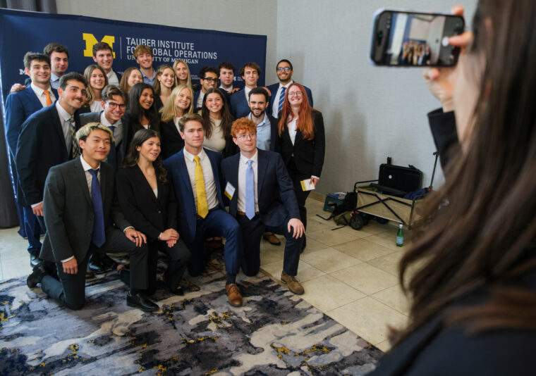 A group of professionally dressed individuals poses for a photo in front of a banner for the Tauber Institute for Global Operations at the University of Michigan, while someone captures the moment with a smartphone.