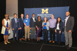 A group of nine individuals stands on a stage in front of a banner for the Tauber Institute for Global Operations at the University of Michigan, with one person holding an award.