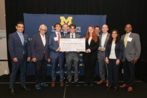 A group of professionals stands together on stage, holding a large ceremonial check presented by the Global Operations Institute at the University of Michigan.