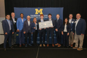 A group of professionals poses with a large ceremonial check in front of a backdrop for the Tauber Institute for Global Operations at the University of Michigan.