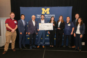 A group of people pose on stage in front of a banner for the Taub Institute for Global Operations at the University of Michigan, with two individuals holding a large ceremonial check.