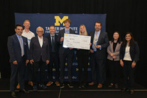 A group of people pose together at an event, with one individual holding a large ceremonial check for $10,000 awarded by the Tauer Institute for Global Operations.